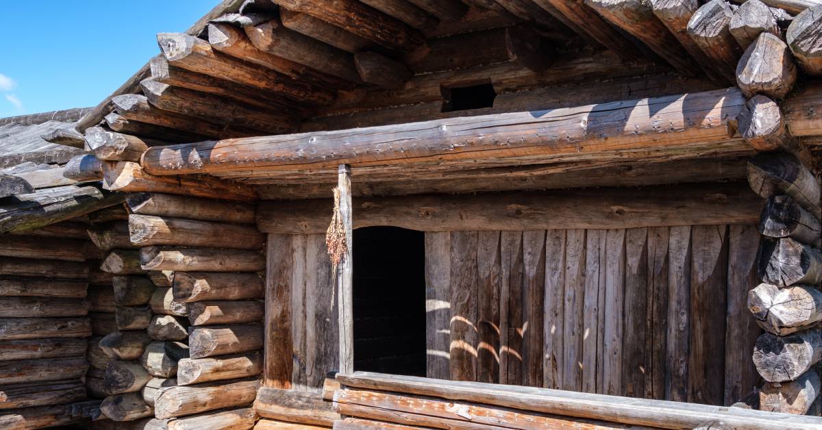 The front of an old log home with discolored wood on it. There’s a porch and the door is open beneath a blue sky.