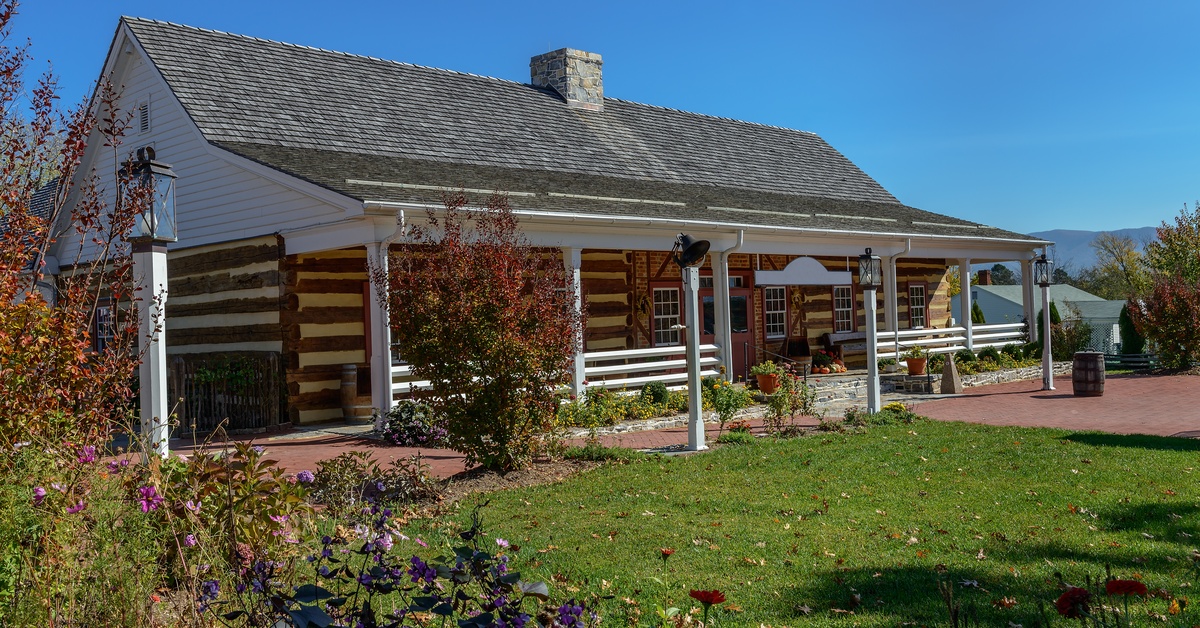A traditional log cabin house with a well-manicured lawn surrounded by bushes and trees beneath a blue sky.