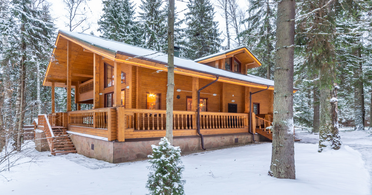 An outside view of a log cabin in the middle of the woods during the wintertime with freshly fallen snow around it.