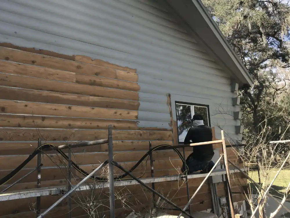 Gray paint partly removed from log siding, showing bare wood beneath, with scaffolding along the wall