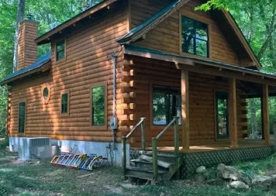 Log home with a stone chimney and green metal roof, ladders staged beside the steps