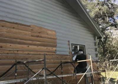 Log siding half stripped of gray paint during restoration, with scaffolding and a crew member at the window